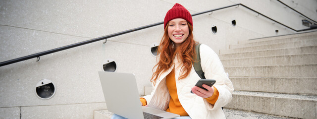 Young tourist, girl booking hotel room on laptop, enters confirmation code on smartphone, sitting with computer and phone on stairs outdoors - obrazy, fototapety, plakaty