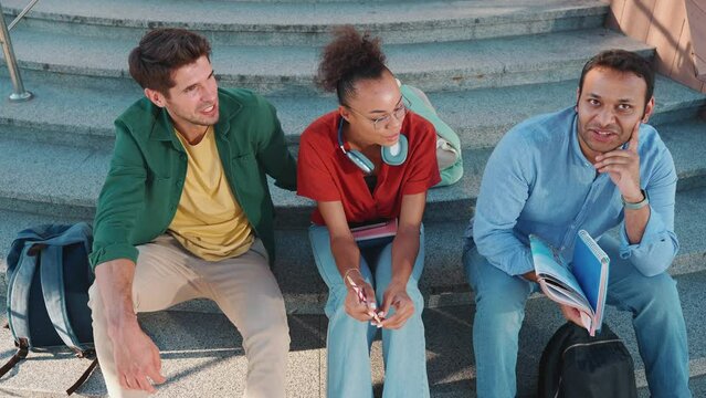 Top view of pretty multiracial students talking while sitting on the stairs outdoors 