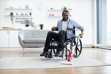 Relaxed adult person in jeans taking lightweight stick vacuum while using wheelchair in spacious flat. Cheerful african homeowner enjoying housework with modern appliances while waiting for guests.