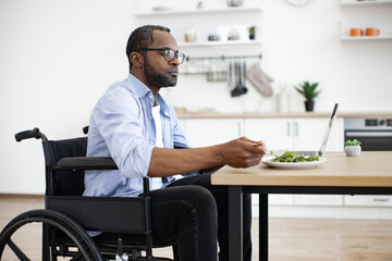 Positive african adult with disability having meal while scrolling webpages on laptop in spacious dining room. Well-organised employee combining lunch break and flexible schedule at distance work.