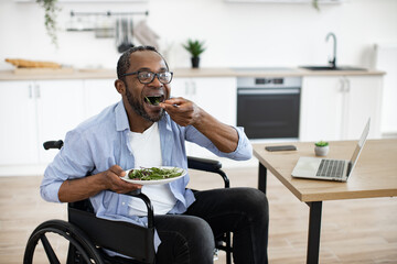 Salad placed on writing desk near laptop by african person with disability in dining room. Happy business manager enjoying healthy food while telecommuting in home office at midday.