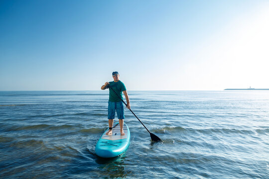 A man in shorts and a T-shirt stands on a SUP board with a paddle near the seashore in the morning at dawn.