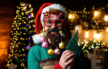 Merry Christmas and Happy New year. Bearded man in Santa hat and party glasses with small Christmas tree. Smiling man with decorated beard and dyed hair holds little fir-tree. Christmas beard style.