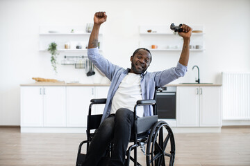 Joyful young adult with disability holding hands with joypad high in air in modern apartment. Smiling african man inishing computer game with winner's gesture indoors.