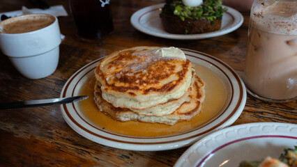 Photographie de pancakes posés dans une assiette blanche, beurre fondu, avocado toast et café 