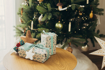 Merry Christmas! Stylish christmas gifts wrapped in festive paper with bows,vintage ribbons, toys on wooden table  against christmas tree lights. Atmospheric winter holidays
