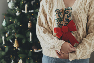 Hands in cozy sweater holding stylish christmas gift in festive wrapping paper with bow on...