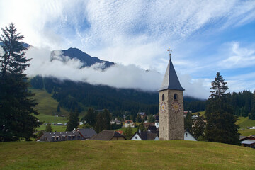 Fototapeta na wymiar Beautiful swiss alpine countryside with a medieval bell tower with a clock and Rothorn mountain on background in Churwalden village in Switzerland formerly Parpan