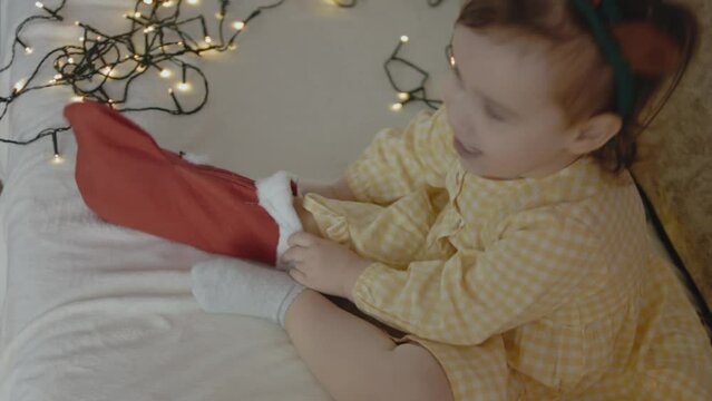 Cute toddler girl playing with her Christmas stocking. She puts on the Christmas stocking on her foot.