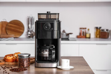 Modern coffee machine with jar of roasted beans, cup and buns on table in kitchen