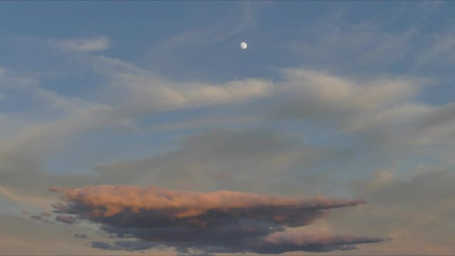 Blue sky, moving beige, orange, pink and blue clouds and silver moon on evening time during sunset - timelapse. Topics: weather, atmosphere, space, dusk
