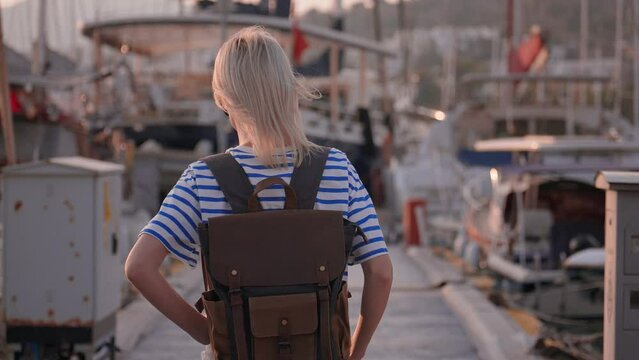 Young beautiful slender blonde woman with backpack in sunglasses, walking on pier against background of sailing ships and yachts, traveling on sea, cruise on ship. Adventure Sunset Travel.