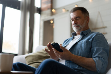 Obraz na płótnie Canvas Happy smiling senior man using smartphone device while sitting on sofa at home