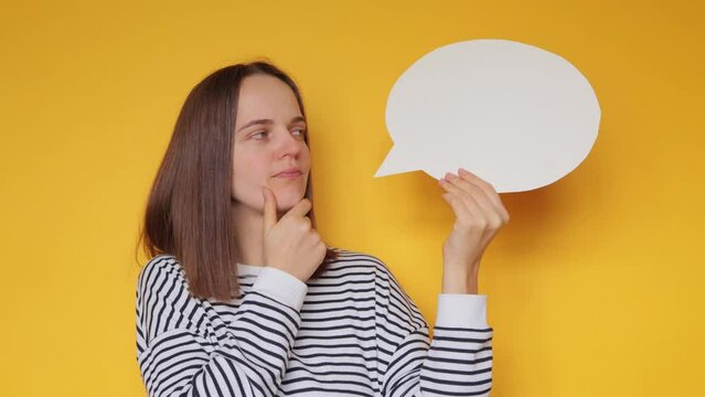 Pensive woman wearing striped shirt holding speech bubble with copy space isolated over yellow background holding chin thinking sharing her thoughts with people.