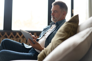 Cheerful senior man in casual clothing using digital tablet while sitting on the sofa at home. - obrazy, fototapety, plakaty