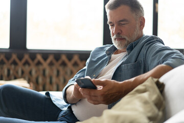 Happy smiling senior man using smartphone device while sitting on sofa at home