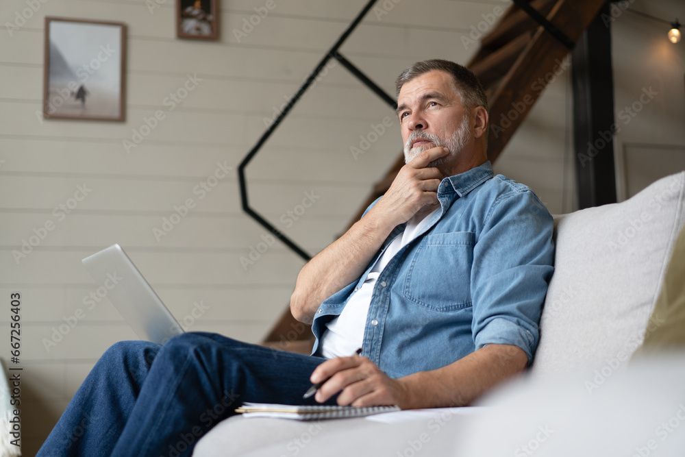 Wall mural Senior man in casual clothing using laptop and smiling while sitting on the sofa, working from home.