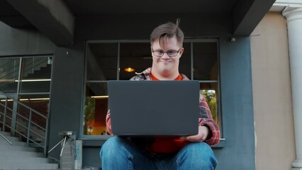 Young man with down syndrome in checkered shirt using laptop working online outdoors in the city. Guy with disability using laptop to make remote video call