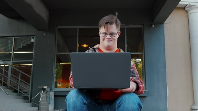 Young man with down syndrome in checkered shirt using laptop working online outdoors in the city. Guy with disability using laptop to make remote video call