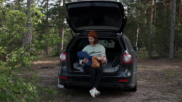 Young Woman Mother 30 Years Old In Hiking Clothes Sits On The Trunk Of A Car With A Child In Her Arms, Breastfeeds And Looks At The Sea, Road Trip, Tourism With A Child