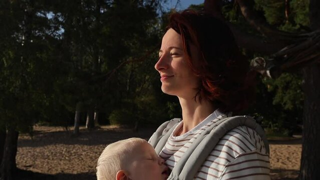 A 30-year-old brown-haired woman stands with her eyes closed with the rays of the sun on her face, enjoying nature, the child is sleeping in an ergo backpack