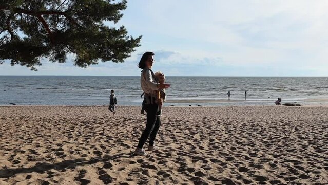 a young woman mother walks along the beach towards the sea, beckons and waves her hand, view from the back