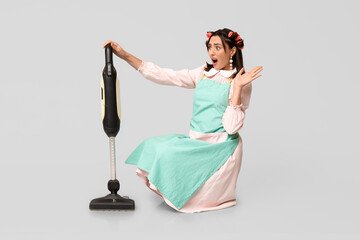 Portrait of shocked young housewife in apron with vacuum cleaner on grey background