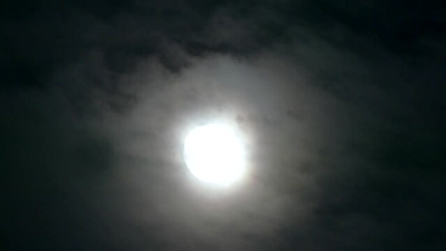 Dark night sky and silver moon with glow around on moving clouds - real time with zoom out. Topics: astronomy, optical phenomenon, atmosphere, cosmos, science, space, nighttime
