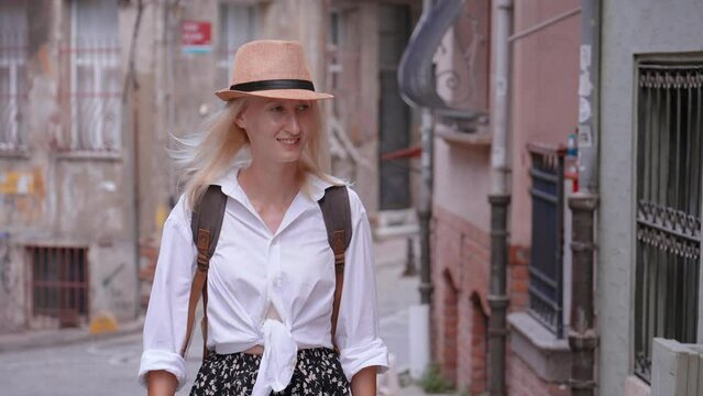 Portrait of caucasian woman beautiful tourist woman with backpack in white shirt and straw hat walking along empty street in old tourist part of city, enjoying her vacation. Sightseeing in popular tou
