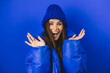 Optimistic woman raises palms from joy, happy to receive awesome present from someone, shouts loudly, dressed in blue puffer jacket, hat, isolated on blue background. Excited female yells, raised hand