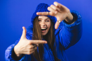 Attractive woman with a lovely smile making a frame gesture with her fingers framing her face. Woman posing on selfie photo looking at camera standing isolated blue background. Girl wear puffer, hat.