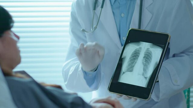 Doctor in gloves showing chest x-ray on digital tablet and zooming lung image for female patient lying on bed in hospital ward. Rack focus shot