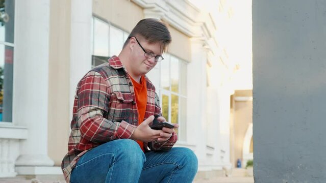 Young man with down syndrome in checkered shirt using phone outdoors in the city. Guy uses smartphone to read news