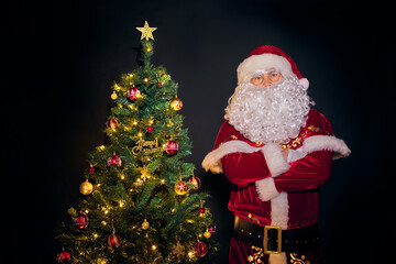 Santa Claus stands next to a beautifully decorated and illuminated Christmas tree on a black background. Xmas decoration and illumination concept.