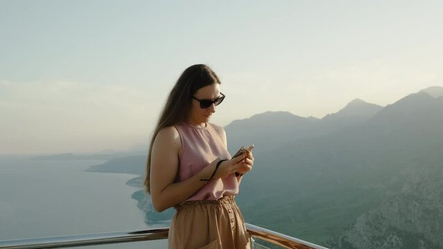 A young woman shares photos of her journey on social media while standing high in the mountains with the sea and sunset in the background.
