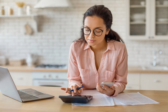 Focused woman with laptop, cellphone and calculator in kitchen