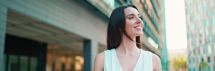 Lovely young woman with freckles and dark loose hair wearing white top and jeans is enjoying the view of modern city. Beautiful girl with long eyelashes looks at the upper floors of building