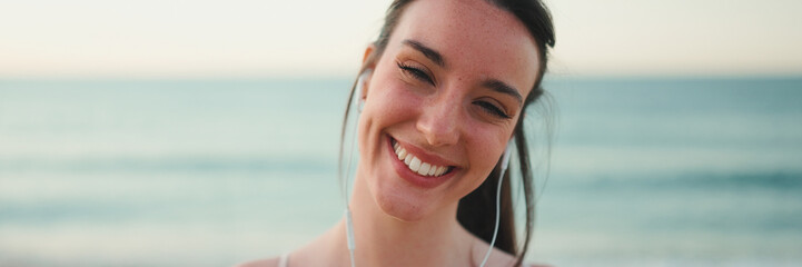 Young athletic woman with long ponytail wearing beige sports top in wired headphones, smiling at the camera while standing on the sea background