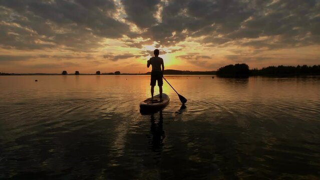 Siluet of man standing firmly on inflatable SUP board and paddling through shining water surface. Wide shot sport man at sunsat on a lake