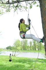 An athletic girl climbs a tall tree using a carabiner belay, pulls herself up onto the rope with her hands, and gains great height. Recreation and sports.