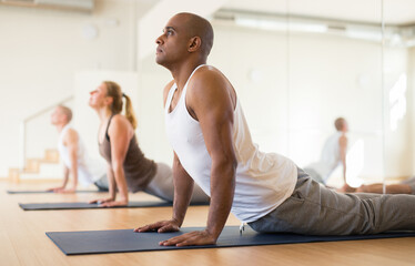 Obraz premium Hispanic man practicing yoga postures during group training at gym, performing stretching asana Urdhva Mukha Shvanasana (Upward Facing Dog Pose)
