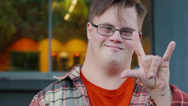 Portrait of young man with down syndrome in checkered shirt outdoors in the city showing thumbs up gesture. Man with trisomy 21 illness. Disabled guy having fun