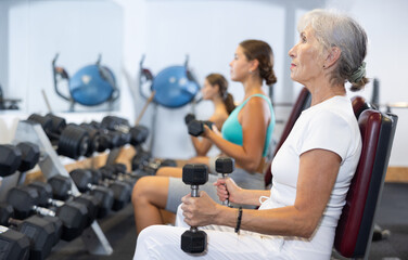 Slender energetic athletic senior woman sit and trains muscles of arms and back using dumb-bell...