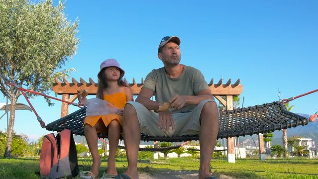 Family eating in hammock. A happy child with her dad relax in the hammock and eat bun against green sunny sky.