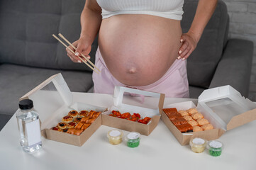 A pregnant woman holds chopsticks and stands at the table with rolls. Close-up of the belly.