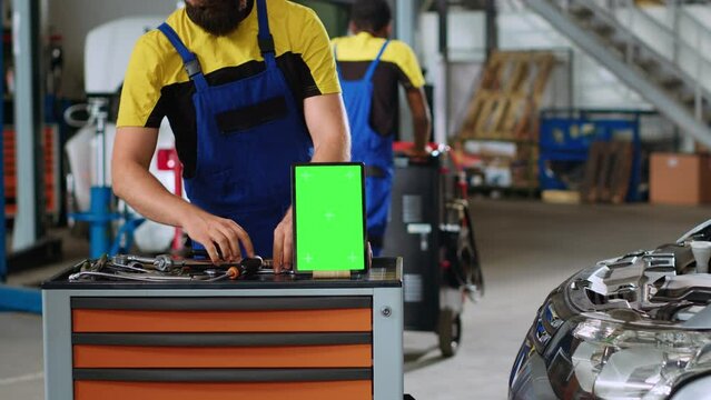 Panning shot of chroma key tablet in garage workspace sitting on work bench. Mockup device in car service next to professional tools with repairmen in background refurbishing damaged vehicles