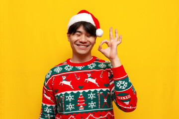 young asian man in christmas sweater and santa hat smiling and showing ok gesture on yellow...