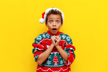 shocked african american boy in christmas sweater and santa hat looking surprised on yellow isolated background