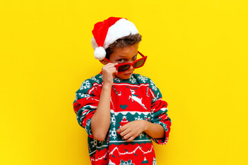 african american boy in christmas sweater and santa hat looks through glasses on yellow isolated...