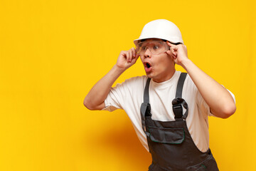 shocked asian male construction worker in uniform and safety glasses looking in amazement over yellow background - obrazy, fototapety, plakaty